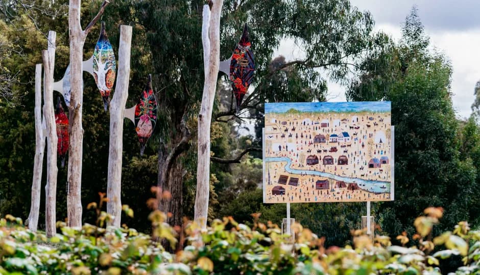 Outdoor art installation featuring three colorful glass lanterns hanging from white, branch-like structures and a square painting depicting a village or landscape on a clear stand, surrounded by greenery.
