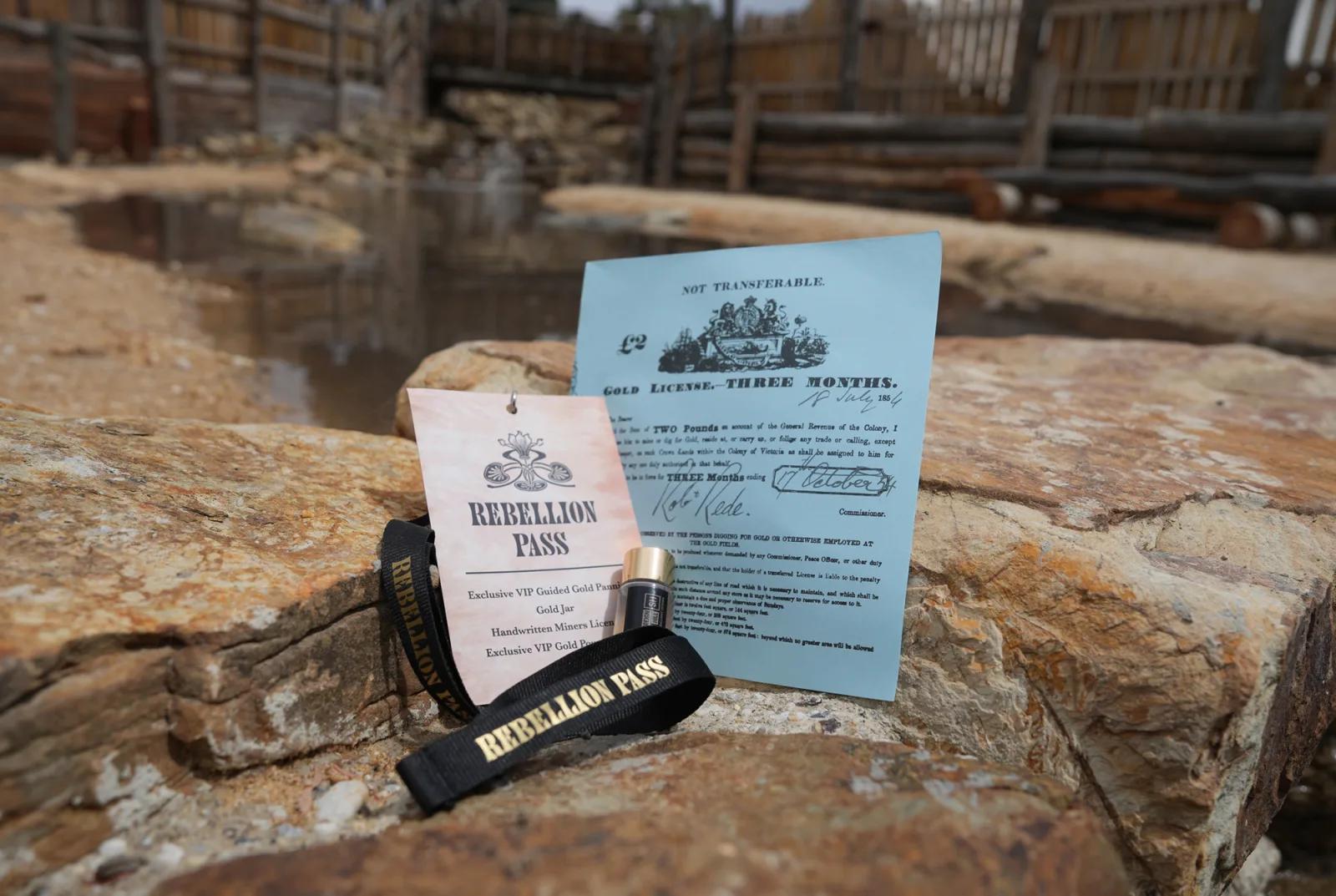 A “Rebellion Pass” with a lanyard and a blue informational flyer are placed on a large rock near a muddy, fenced outdoor area. The pass and flyer suggest entry to a historical or themed attraction.