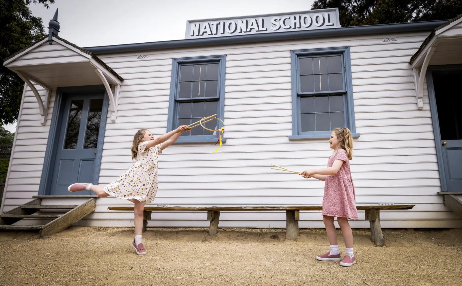 Two young girls play joyfully with wooden bats outside the historic National School building at Sovereign Hill, capturing the charm of 19th-century childhood and interactive learning experiences offered at Sovereign Hill’s Games National School program.