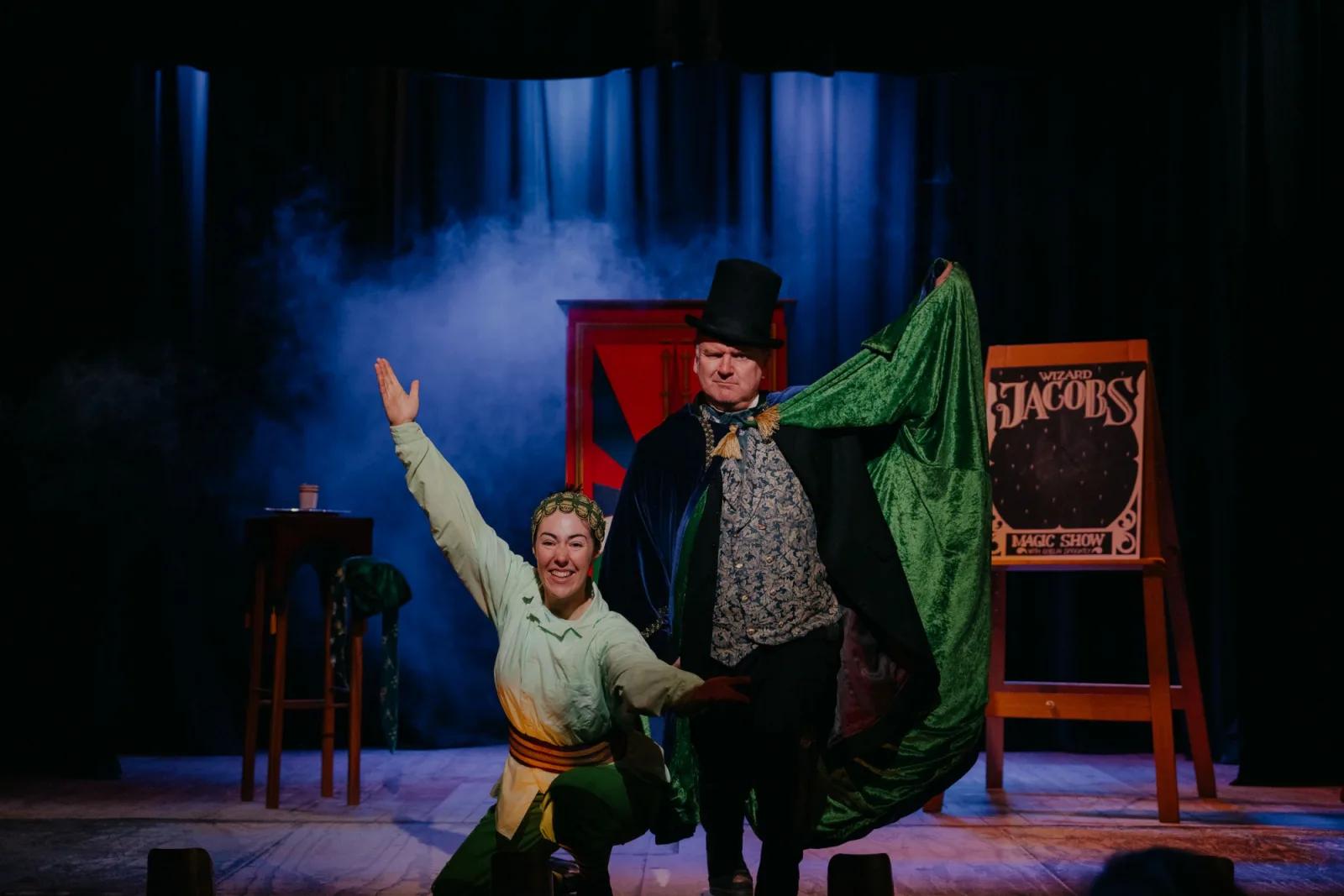 On stage at Sovereign Hill, performers from The Common Wanderer bring a theatrical gold rush tale to life, one actor in a top hat and green cape gestures dramatically beside another mid-gesture, set against moody blue lighting and vintage props. A captivating blend of history and storytelling at Sovereign Hill.