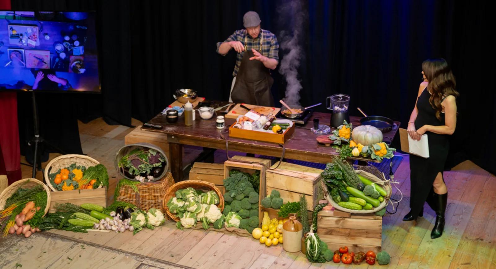 At Sovereign Hill, a dynamic live cooking demonstration unfolds on stage, where a chef in a plaid shirt and apron prepares a dish amid an abundant display of fresh produce, including carrots, broccoli, pumpkins, and leafy greens, arranged in wooden crates and woven baskets. A host in a black dress observes with a clipboard, while a screen projects the action, capturing the interactive and immersive culinary experience that brings heritage-inspired cooking to life at Sovereign Hill.