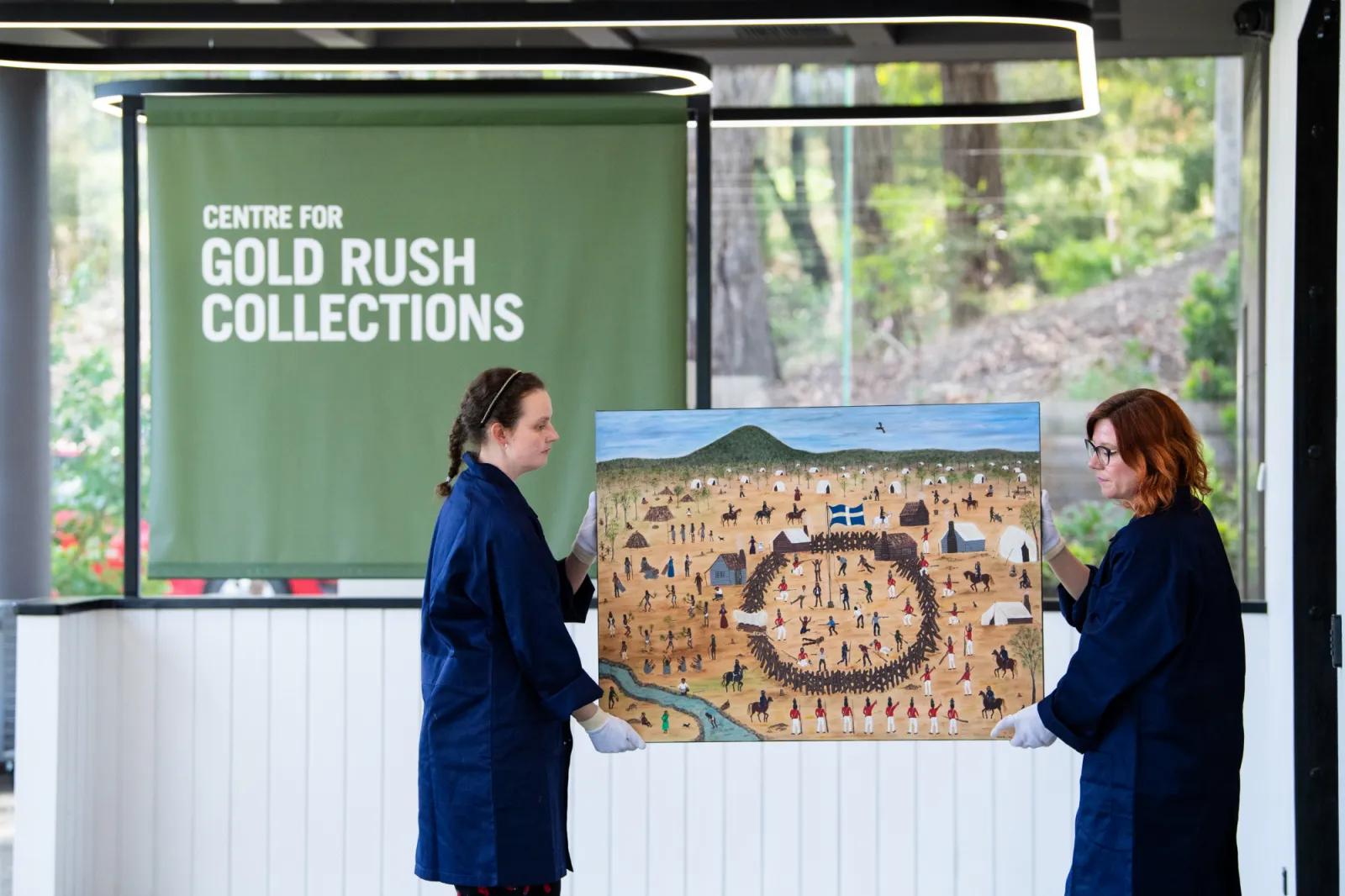 Two people in dark blue smocks and white gloves carefully holding a large, colorful painting depicting a historical scene with numerous figures, horses, and flags, in front of a green banner that reads 'CENTRE FOR GOLD RUSH COLLECTIONS'.
