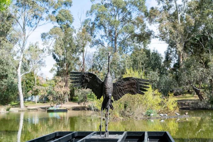 A black metal crane sculpture with outstretched wings standing on a platform in a pond, surrounded by trees and greenery.