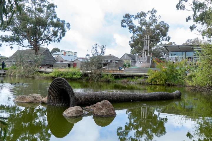 A large, woven, funnel-shaped sculpture partially submerged in a pond, with rocks at its base and a wooden walkway and buildings visible in the background.