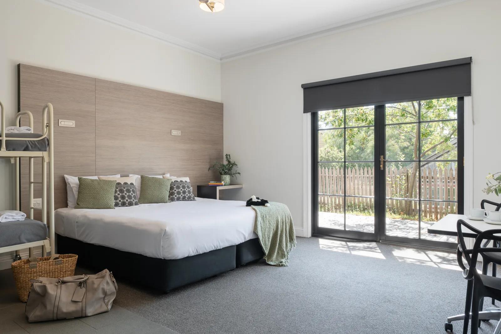 A bright, modern Sovereign Hill Hotel Superior Family Room features a queen bed with patterned green and black pillows, a bunk bed for children, and a private patio accessible via French doors offering comfort, space, and natural light for families visiting Sovereign Hill.