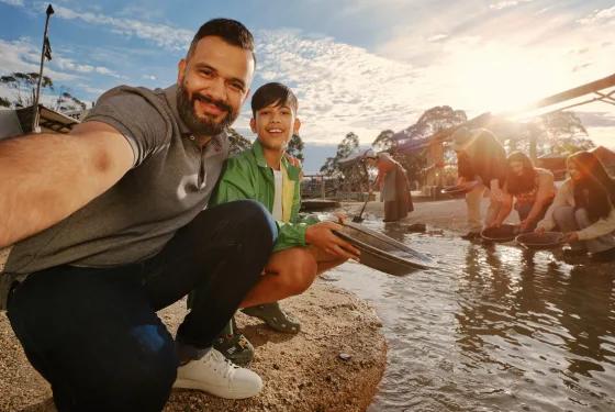 At Sovereign Hill, a man and boy capture a joyful selfie while gold panning along a sunlit creek, surrounded by other visitors engaged in the hands-on activity. Historic gold rush-era buildings line the background under a bright blue sky, highlighting the interactive, family-friendly charm of Sovereign Hill’s living history experience.