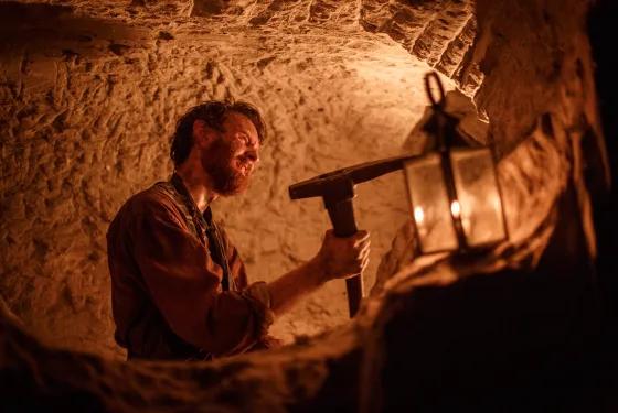 Costumed miner using a hand tool by lantern light inside an underground mine during an applied learning experience at Sovereign Hill.